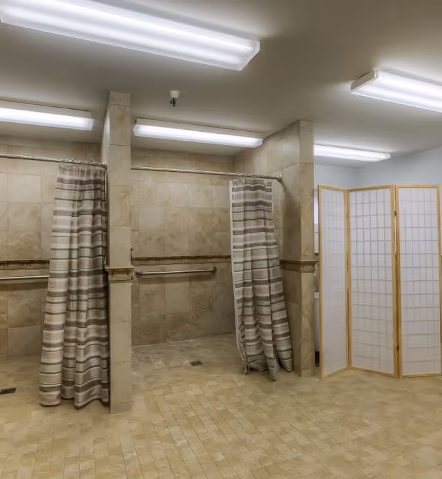 Interior view of a senior living facility bathroom with two shower stalls separated by a tiled wall, each with a striped shower curtain. There are grab bars on the walls inside the stalls and a folding privacy screen on the right side. The floor and walls are tiled in beige tones, and fluorescent lights are mounted on the ceiling.