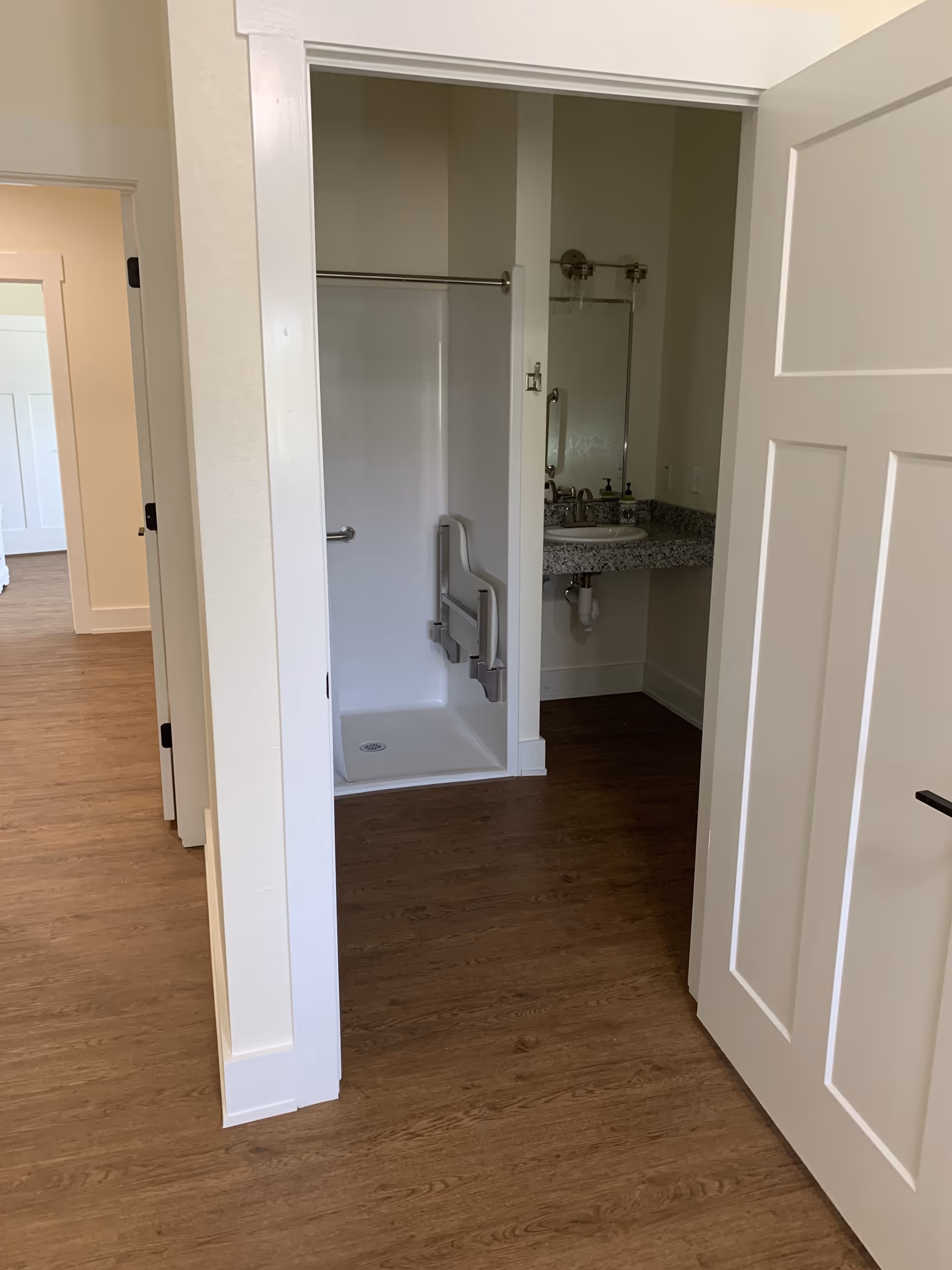 View into a bathroom from an adjacent room showing a walk-in shower with a foldable seat and grab bars, a granite countertop with a sink, a mirror, and a soap dispenser. The floor is wood-style laminate, and the walls are painted light beige.