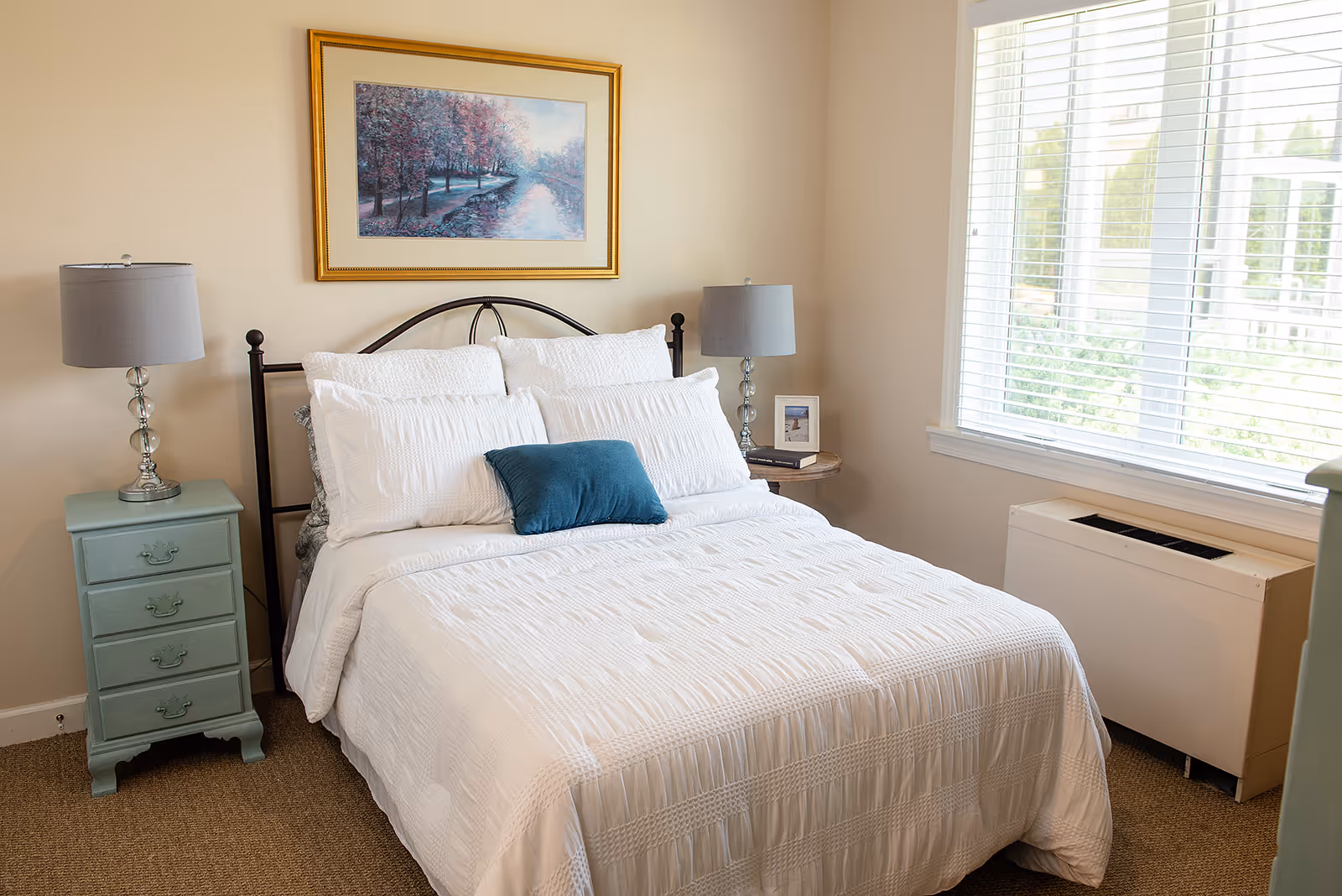 A neatly made bed with white bedding and a teal throw pillow in a sunlit bedroom with bedside tables and lamps.