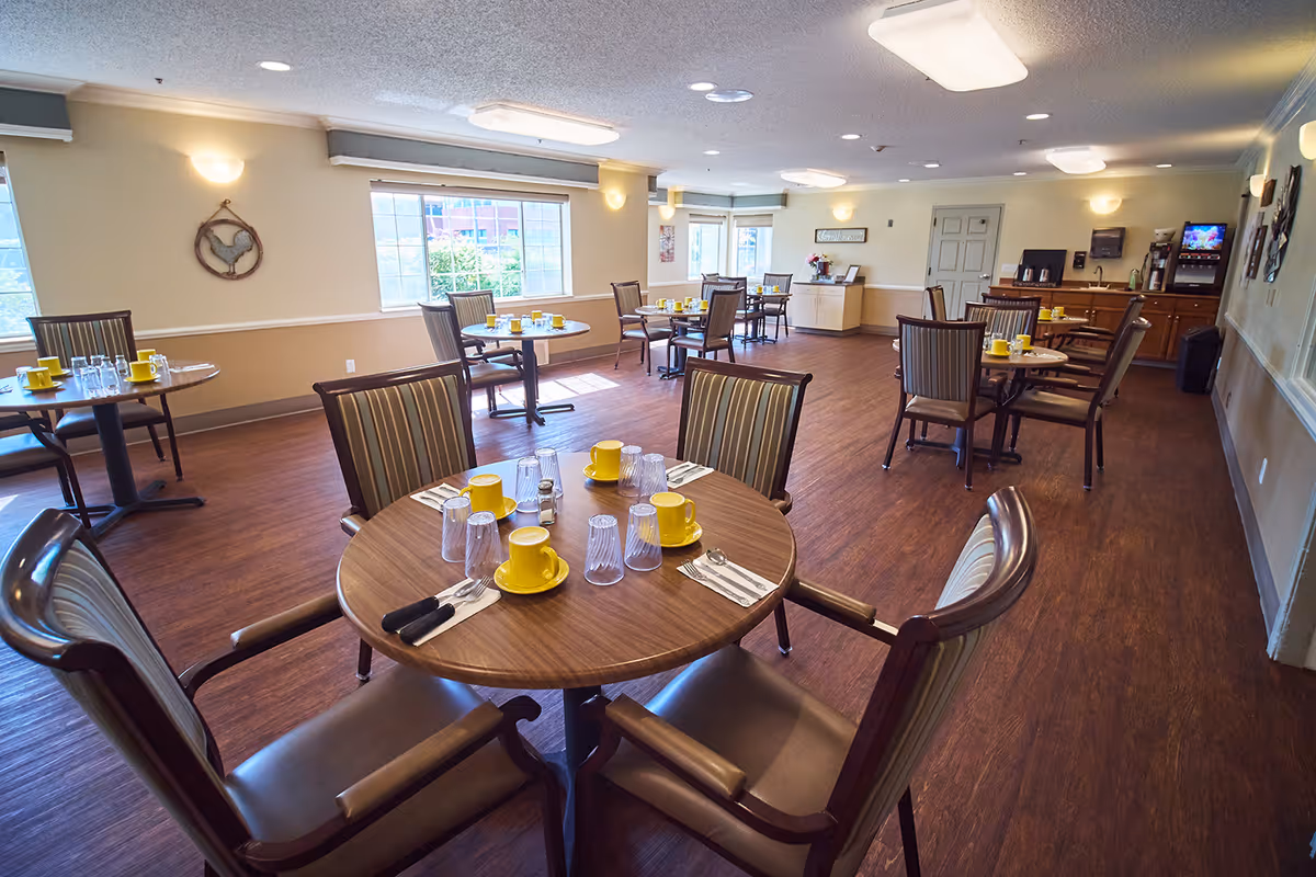 A bright dining room with multiple round wooden tables set with yellow cups, glasses, and silverware. The room has wooden flooring, beige walls with white trim, and several windows letting in natural light. There are striped cushioned chairs around each table and a beverage station in the back corner.