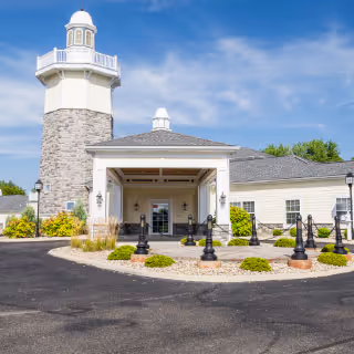 Exterior view of a senior living facility named Altercare Thornville featuring a building with a lighthouse-style tower, a covered entrance, and landscaped surroundings under a blue sky with some clouds.