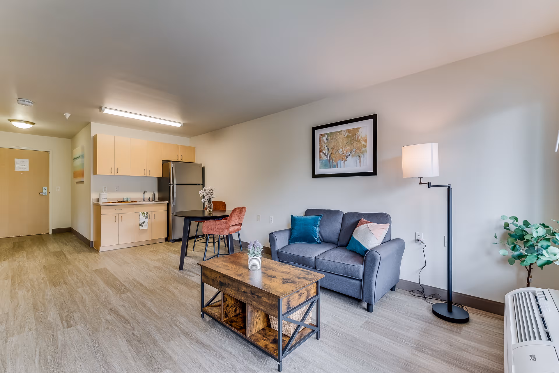 A cozy senior living apartment interior featuring a small kitchen area with light wood cabinets and a stainless steel refrigerator, a round dining table with two red chairs, a gray loveseat with colorful pillows, a wooden coffee table with a small plant, a floor lamp, and a framed artwork on the wall. The room has light-colored wood flooring and neutral walls.