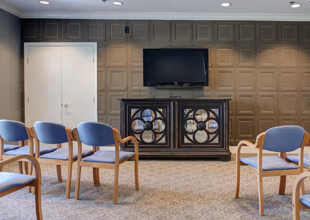 A room with several wooden chairs with blue cushions arranged facing a dark wooden cabinet with decorative circular patterns. Above the cabinet is a flat-screen TV mounted on a wall with a textured panel design. There is a closed white double door on the left side of the wall.