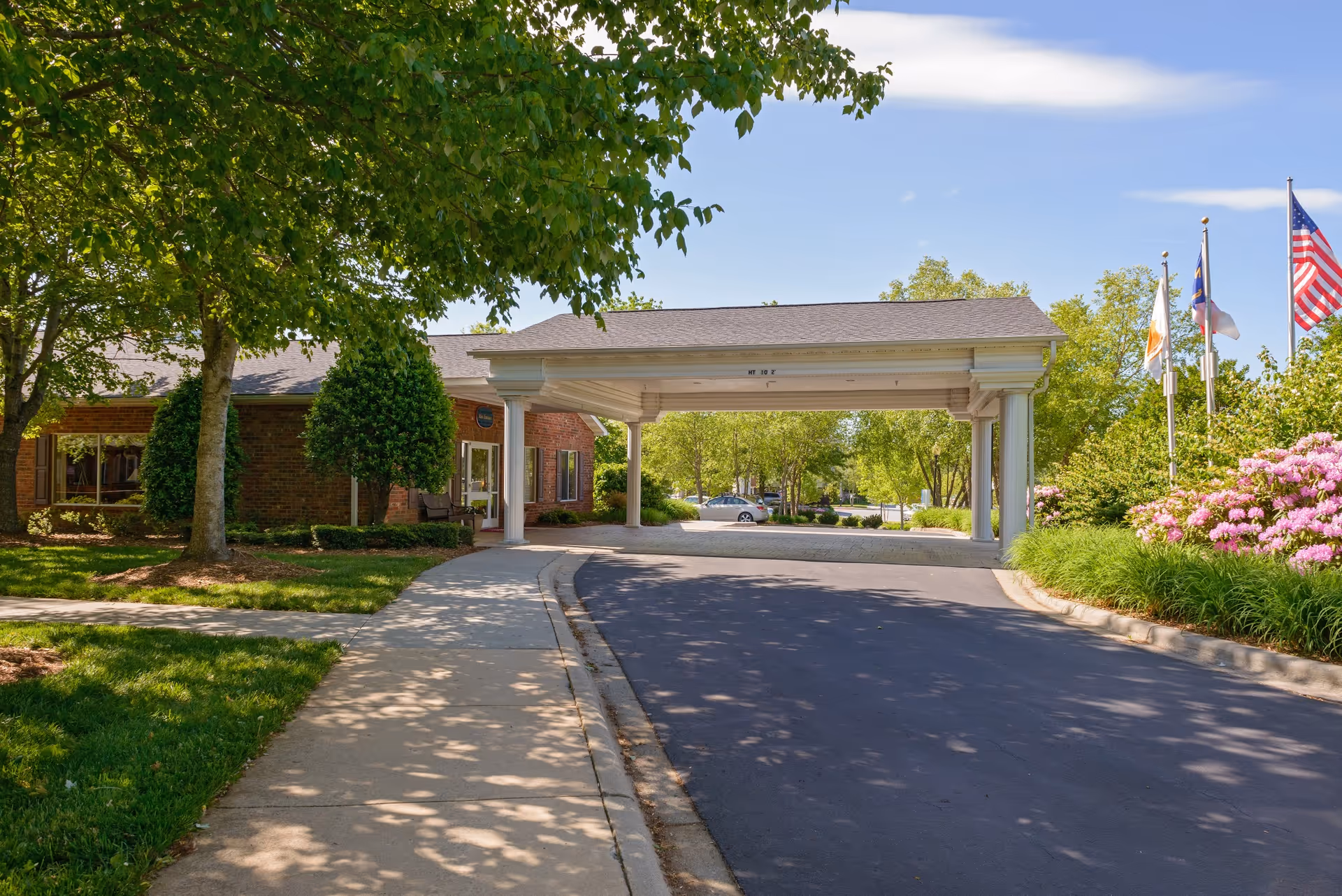 Entrance driveway to a senior living facility with a covered drop-off area supported by white columns. The building is partially visible with brick walls and windows. There are trees, bushes, and flowering plants along the sidewalk and driveway. Several flags, including the American flag, are flying on flagpoles to the right.