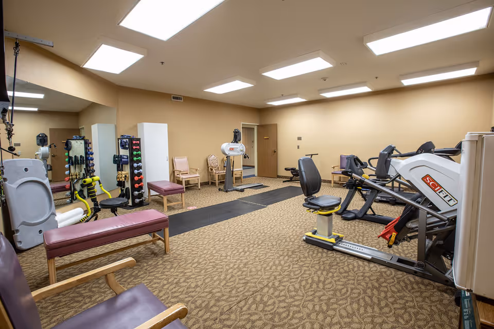 A fitness room in a senior living facility with exercise equipment including a recumbent bike, weight machines, dumbbells on racks, and benches. The room has beige walls, patterned carpet, and bright overhead lighting. Several chairs are placed around the room, and a large mirror covers one wall.