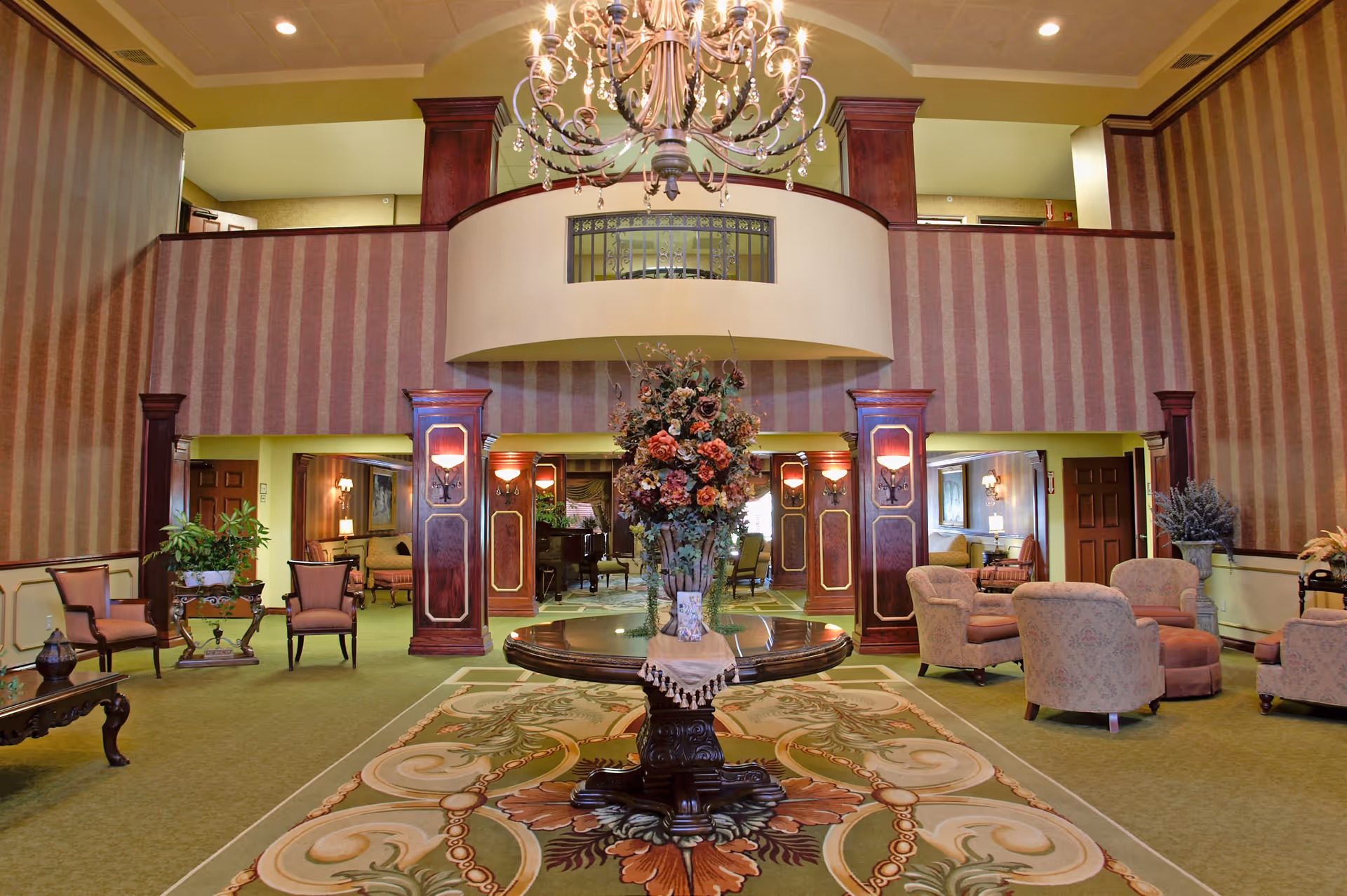 Spacious and elegant living room area with a large floral centerpiece on a round wooden table in the center. The room features striped wallpaper, green carpet, multiple seating areas with armchairs and sofas, wooden pillars with wall sconces, and a grand chandelier hanging from the ceiling. There is a balcony area above with decorative railing.