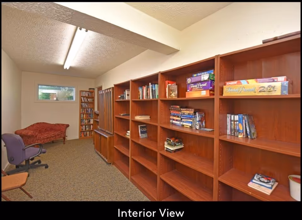 Interior view of a room with wooden bookshelves filled with books and board games, a purple office chair, a red upholstered chaise lounge, and a small window letting in natural light.