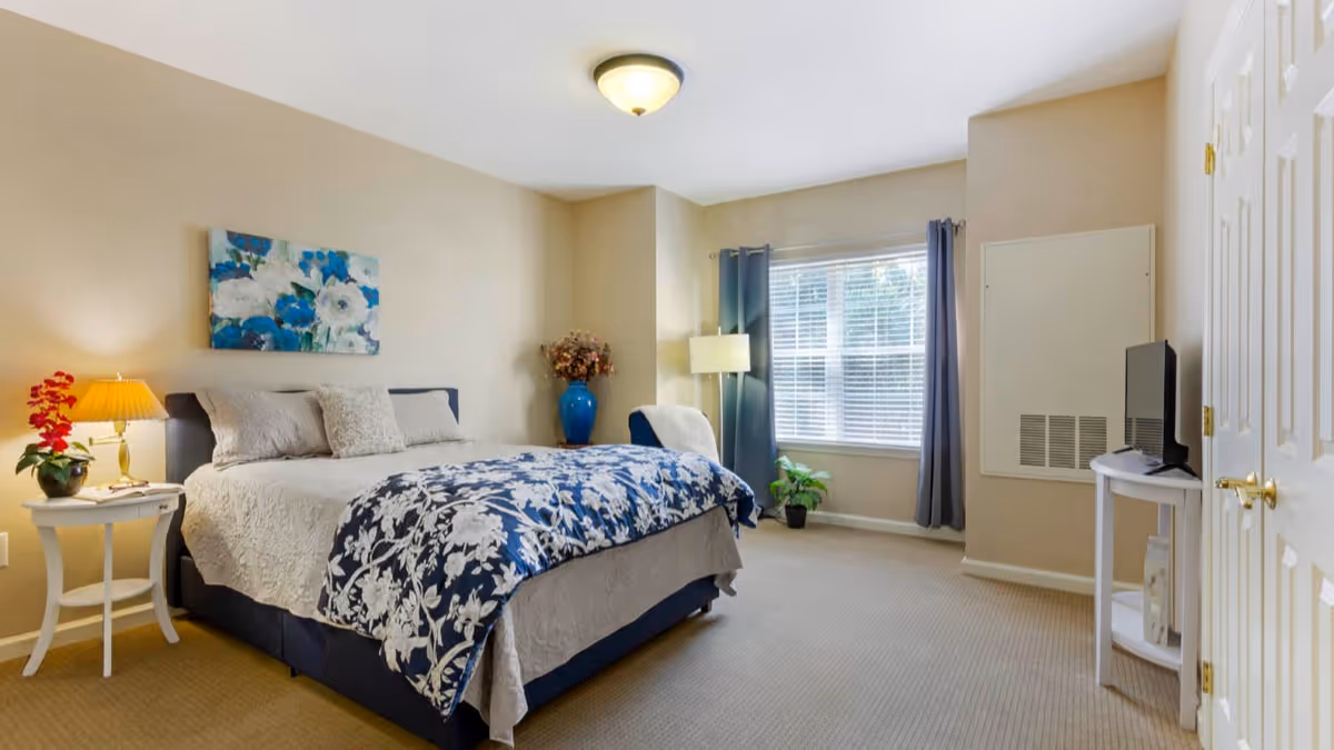 Well-lit bedroom with a bed dressed in blue-and-white floral bedding, bedside tables and lamps, a window with curtains, and a small TV.