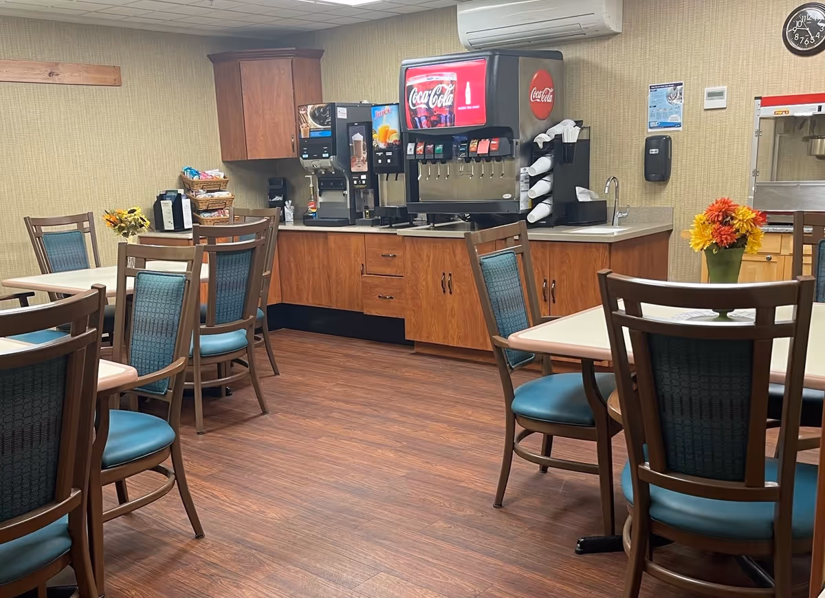 A dining area in Westview Care Center featuring several tables with chairs arranged around them. The room has wood flooring and beige walls. Against the far wall, there is a beverage station with a Coca-Cola soda dispenser, coffee machines, and a popcorn machine. There are small flower arrangements on the tables and a wall clock above the beverage station.