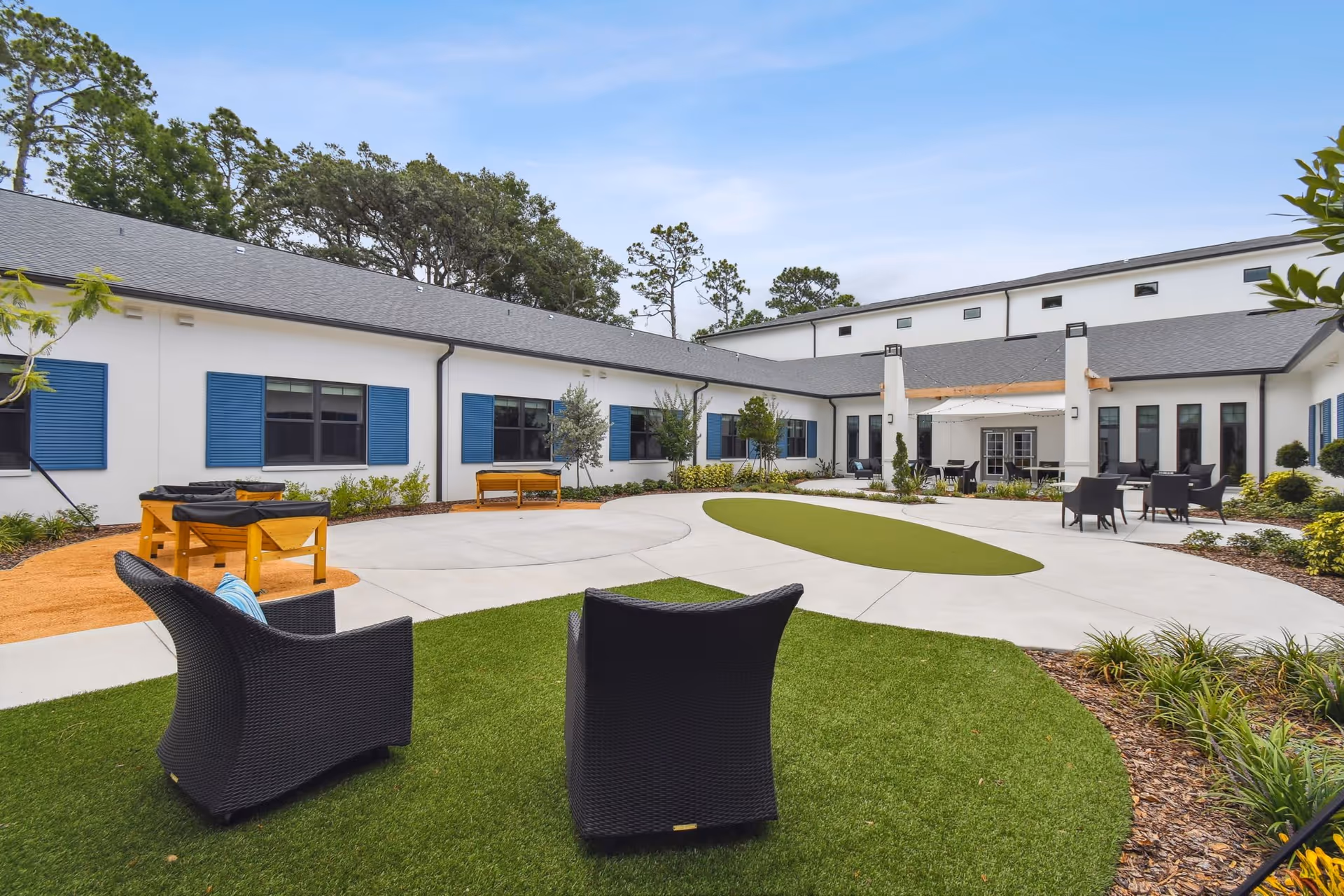 Outdoor courtyard area of a senior living facility with green artificial turf, black wicker chairs, foosball tables, and a white building with blue window shutters surrounding the space under a clear blue sky.