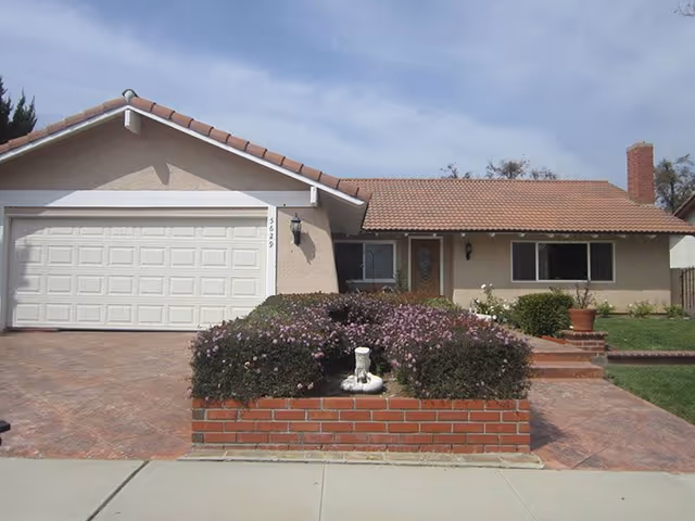 Single-story house with a tiled roof, a two-car garage, a front door with a decorative glass panel, and a brick planter filled with purple flowering bushes in front of the house.