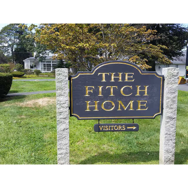 A wooden sign reading "The Fitch Home" with a small "Visitors" arrow standing on a grassy lawn in front of a building.