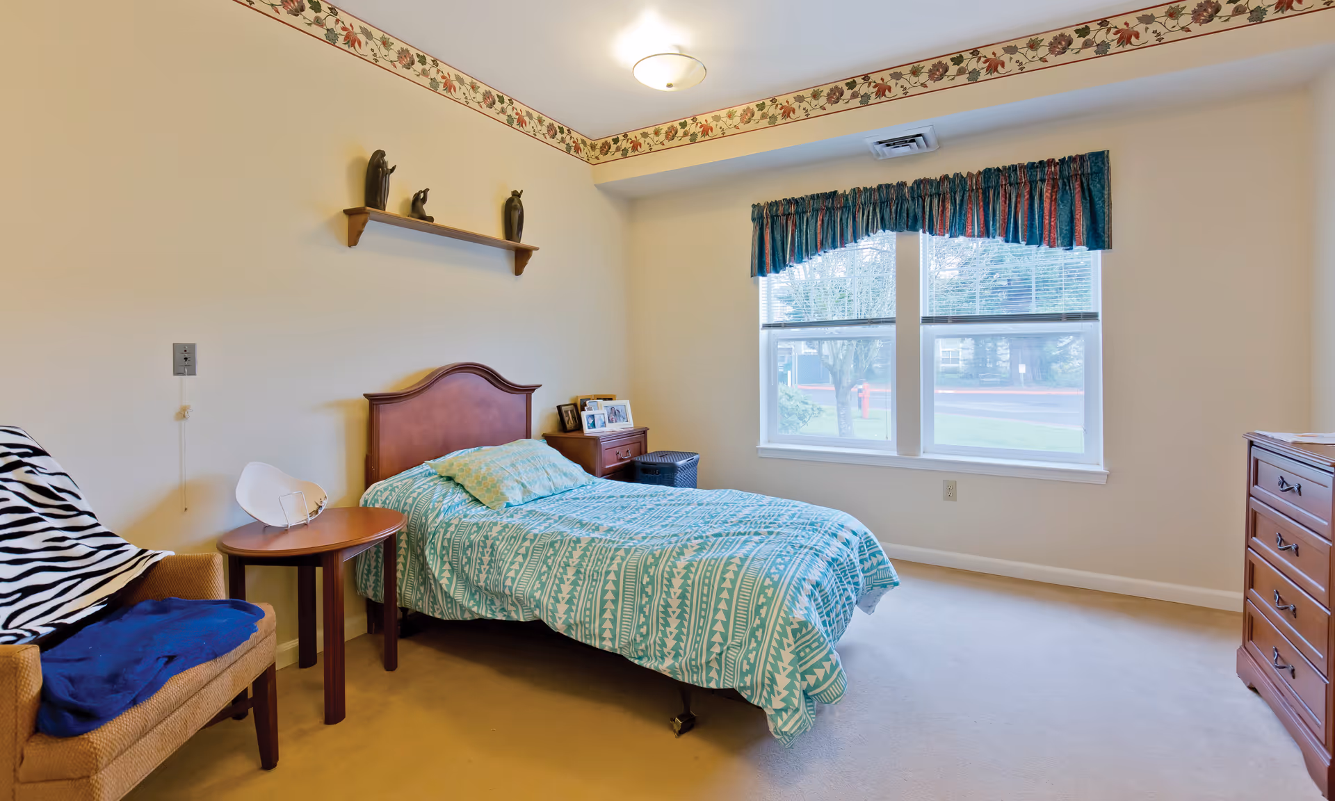A bedroom with a single bed covered in a turquoise patterned bedspread, a wooden headboard, a small wooden nightstand with framed photos, a wooden dresser, a beige armchair with a zebra print blanket and a blue cloth, a small round wooden table, and a window with blue and red valance curtains. The walls are light-colored with a floral border near the ceiling.
