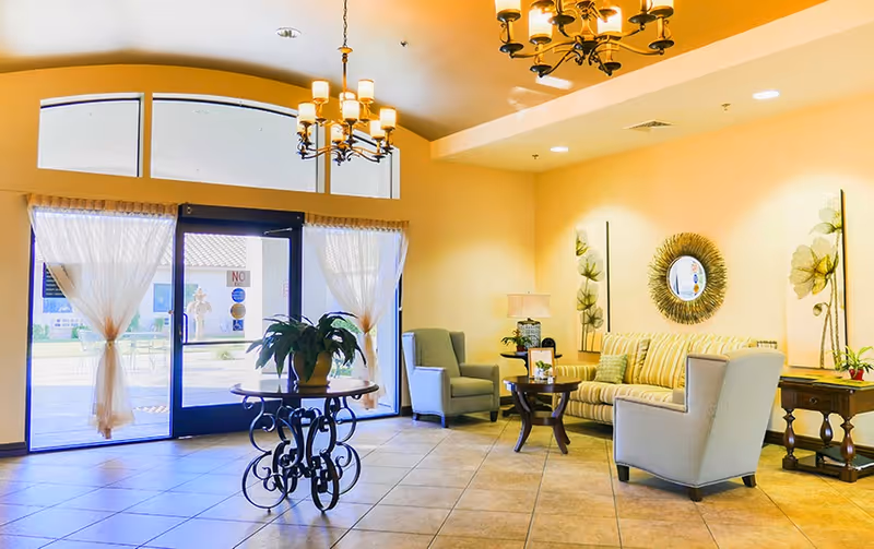 Bright senior living lobby with a sofa, armchairs, decorative tables and chandeliers beside a glass entrance.
