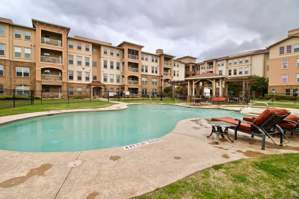 Outdoor swimming pool area with lounge chairs and a covered seating area in front of a multi-story residential building under a cloudy sky.