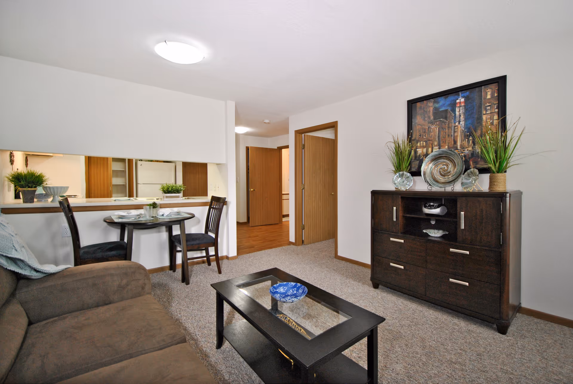 Interior view of a living room area in a senior living facility with a brown couch on the left, a small round dining table with two chairs near a pass-through window to the kitchen, a dark wooden coffee table with a blue decorative bowl, and a dark wooden dresser with decorative plants and artwork above it on the right. Two open wooden doors lead to other rooms.