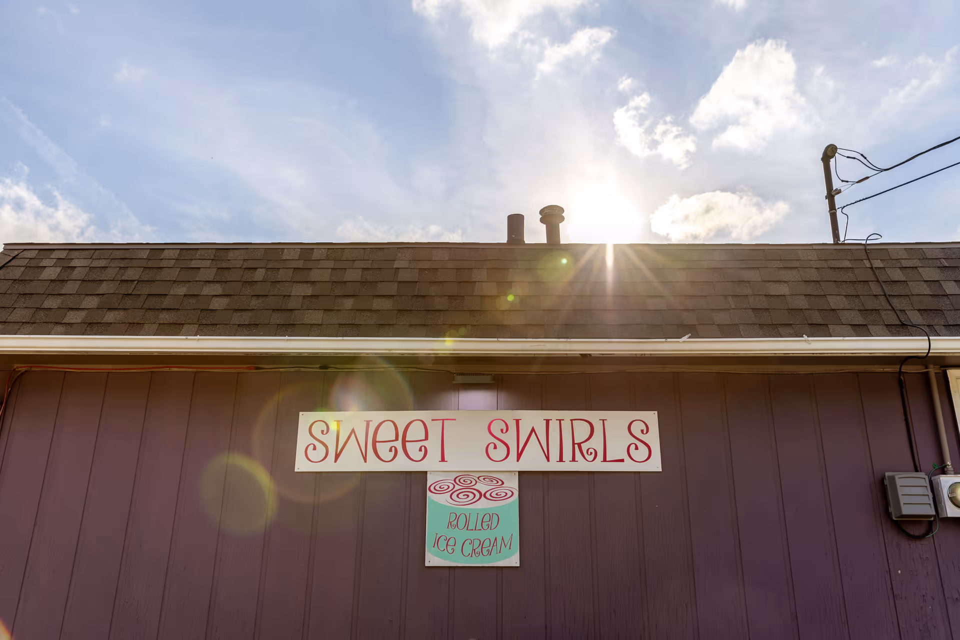 Exterior view of a building wall with a sign that reads 'Sweet Swirls' and a smaller sign below it that says 'Rolled Ice Cream' under a partly cloudy sky with the sun shining brightly above the roof.