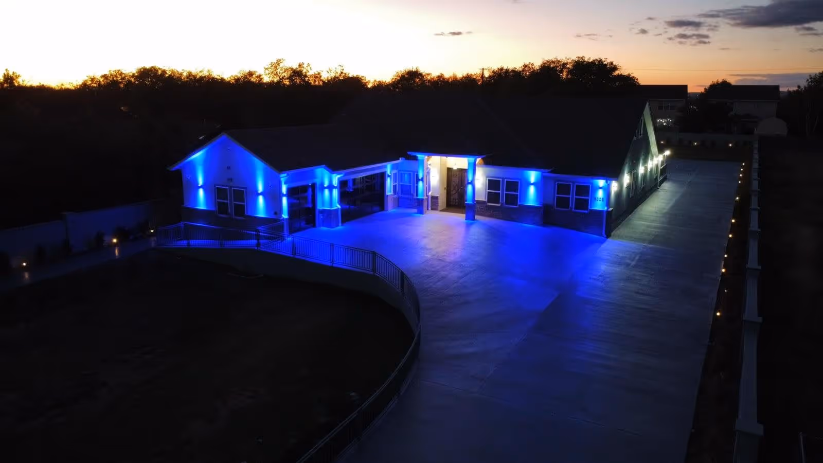 Aerial view of a single-story assisted living facility building illuminated with blue exterior lights at dusk, with a long driveway and surrounding trees in the background.