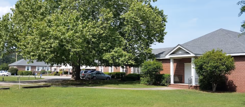 Exterior view of a senior living facility with brick buildings, a large tree, green lawn, and parked cars under a clear blue sky.