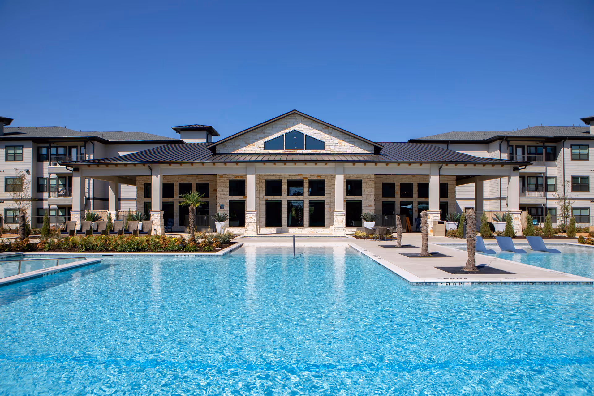 Outdoor view of a senior living facility named Solea Keller featuring a large swimming pool with clear blue water, surrounded by a patio area with lounge chairs and small palm trees. The building in the background has a modern design with stone and light-colored siding, multiple windows, and a covered porch area.