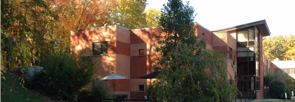 Exterior view of a multi-story brick building surrounded by trees and greenery with outdoor seating and umbrellas visible near the entrance.