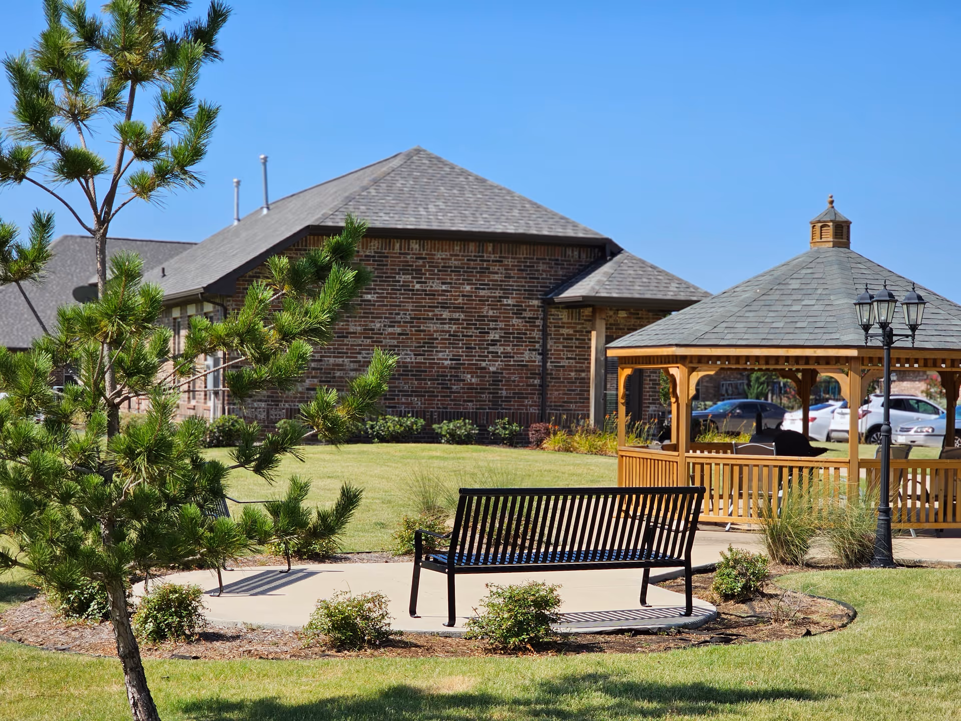 Outdoor garden area at Mayberry Gardens Assisted Living and Memory Care Homes featuring a black metal bench on a concrete pad, a wooden gazebo with a shingled roof, a streetlamp, green grass, small bushes, and a tree with a brick building in the background under a clear blue sky.