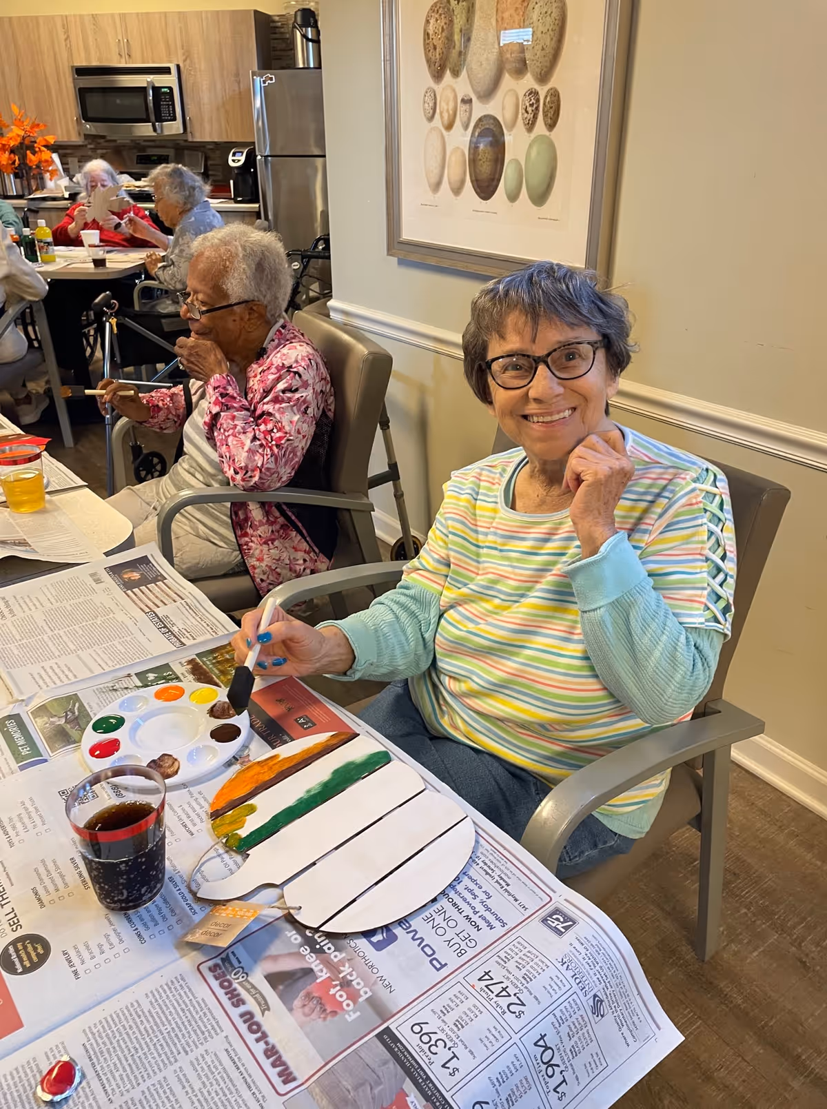 An elderly woman wearing glasses and a striped shirt is smiling at the camera while painting a wooden pumpkin shape at a table covered with newspapers. Other elderly individuals are seated at tables in the background, engaged in various activities. The setting appears to be a communal indoor area with a kitchen visible in the background and a framed picture of various stones on the wall.