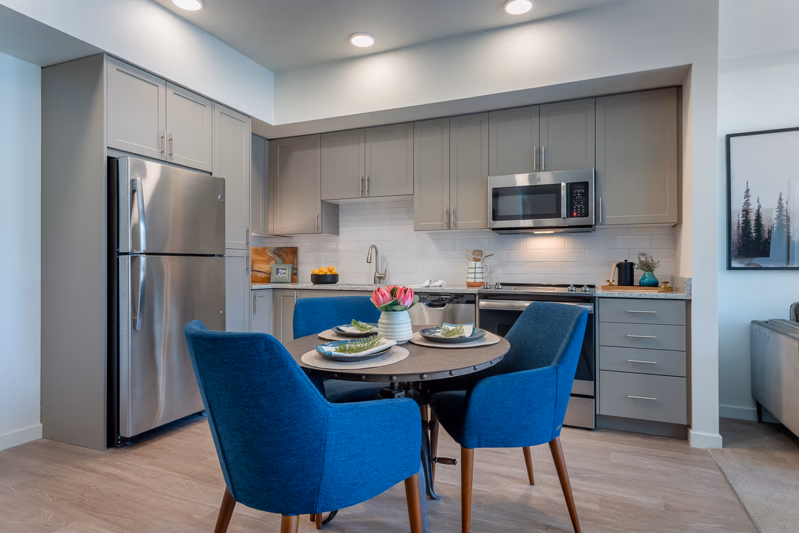 Modern kitchen area with gray cabinets, stainless steel refrigerator, microwave, and stove. A round dining table with four blue upholstered chairs is set with plates, napkins, and a vase of pink flowers. The kitchen has a white subway tile backsplash and light wood flooring.
