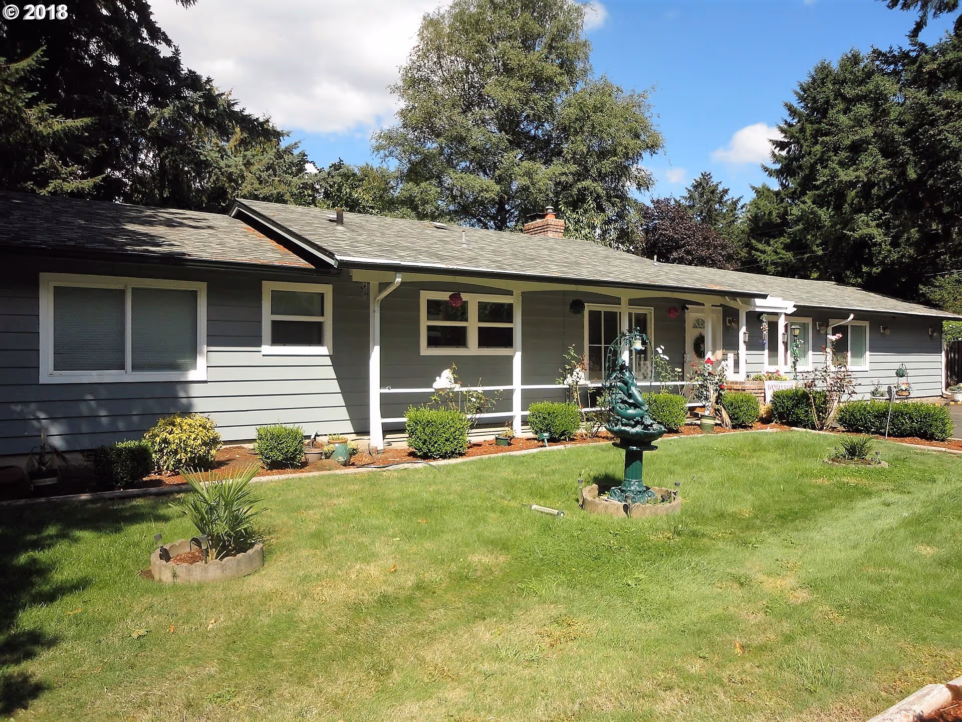 Single-story gray building with white trim surrounded by a well-maintained lawn and garden. There are several bushes and small plants along the front of the building, and a decorative green fountain is centered on the lawn. Tall trees are visible in the background under a partly cloudy sky.