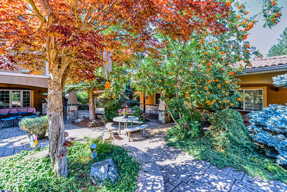 Outdoor courtyard area at Courtyard at Coeur d'Alene featuring a round picnic table with benches, surrounded by lush greenery including trees with red and green leaves, flowering plants, and shrubs. The courtyard is paved with stone tiles and there are buildings with windows and a tiled roof in the background.