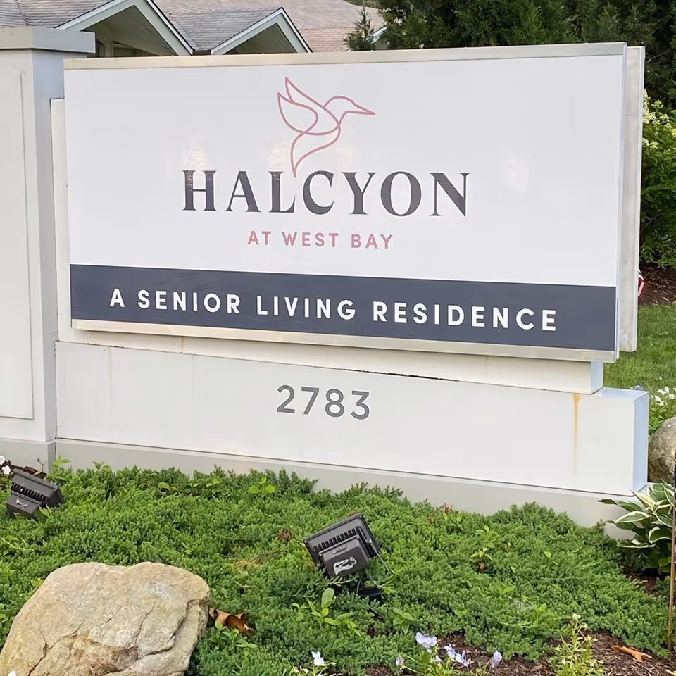 Outdoor sign for Halcyon at West Bay, a senior living residence, with the address number 2783 displayed below. The sign is surrounded by greenery and landscaping rocks.
