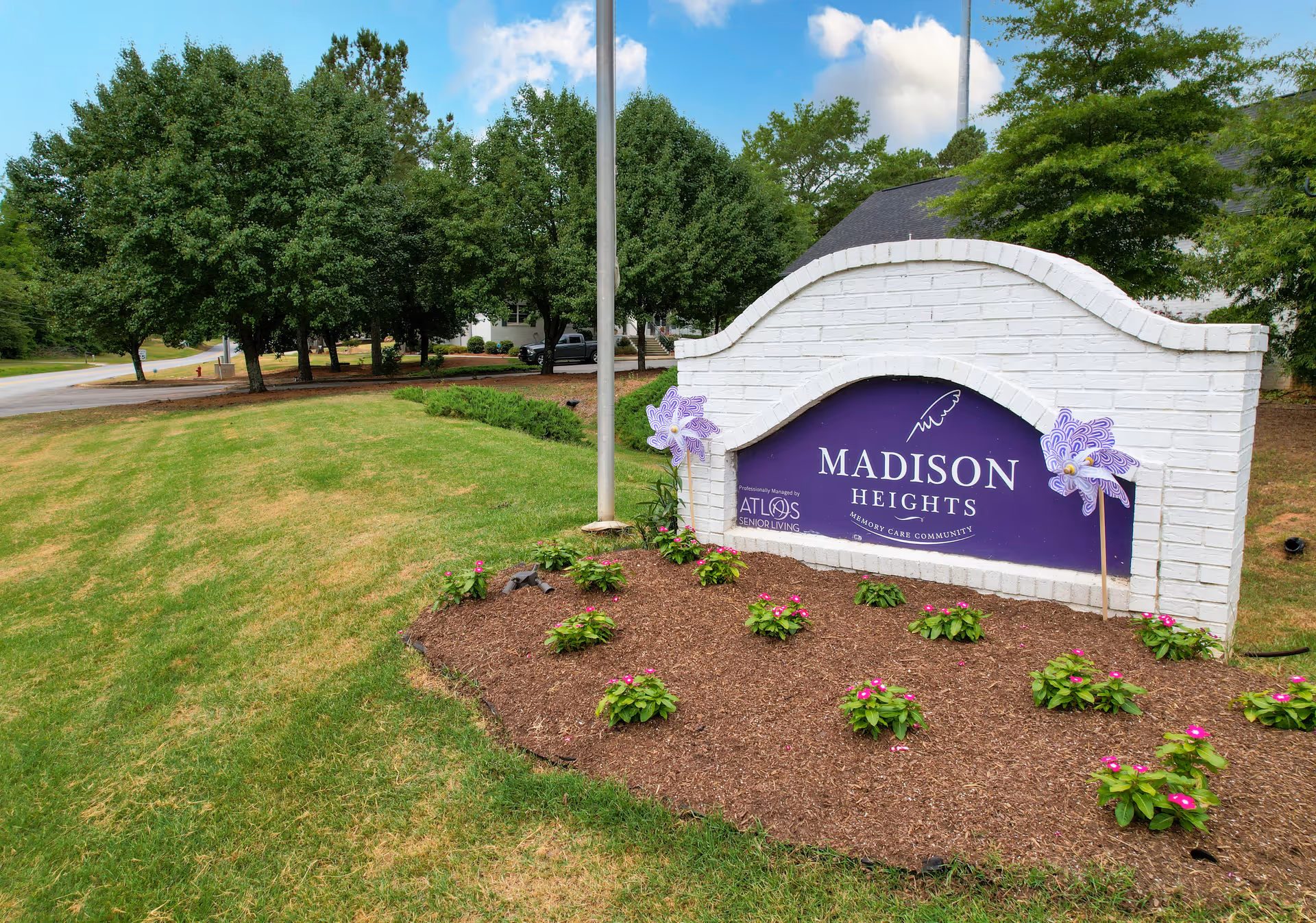 Outdoor view of a landscaped area with a white brick sign displaying 'Madison Heights Memory Care Community' in purple, surrounded by small plants and flowers, with trees and a road in the background under a partly cloudy sky.