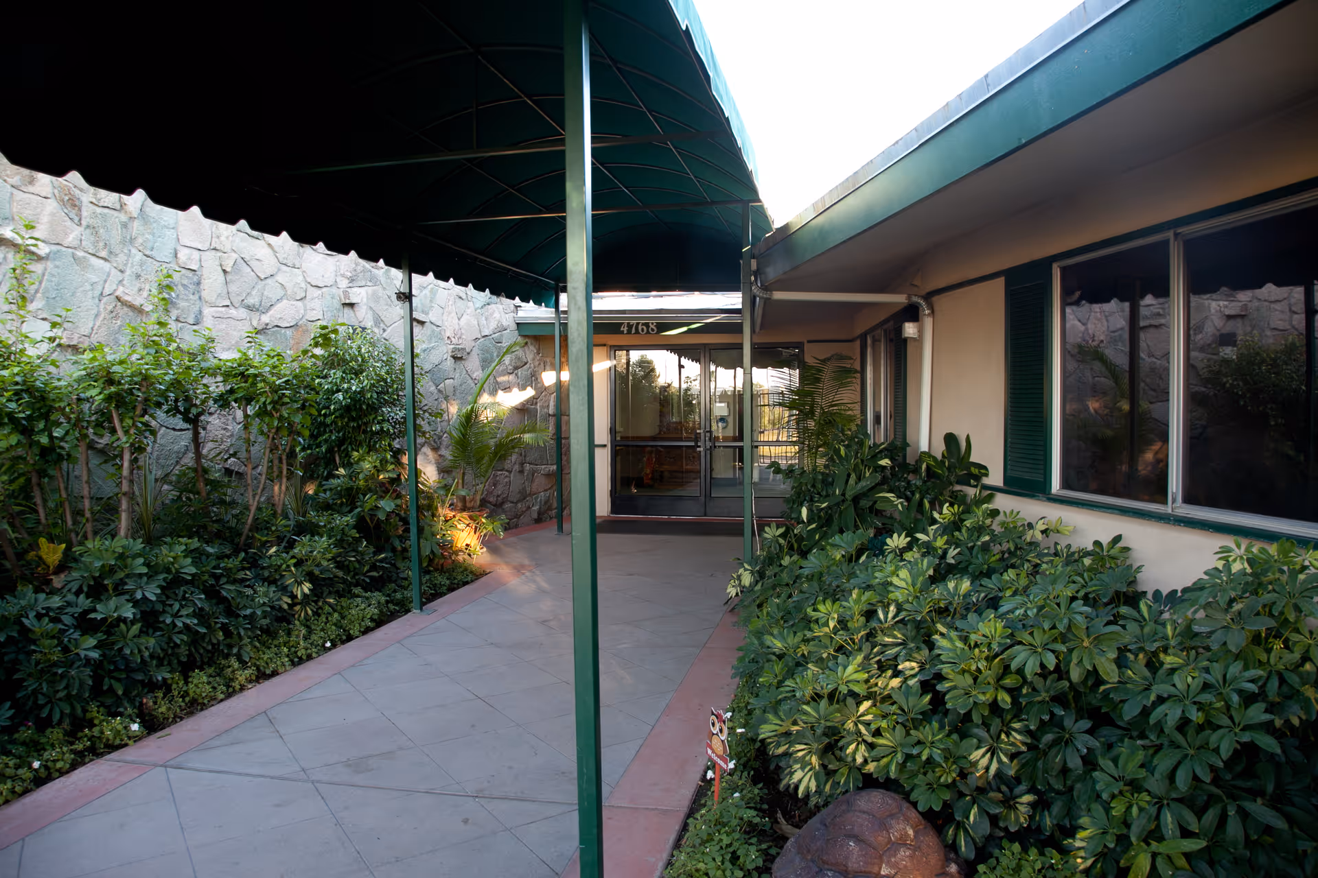 Covered walkway leading to the entrance of a building with glass doors, surrounded by green plants and bushes on both sides. The left side has a stone wall, and the right side shows windows with green shutters.