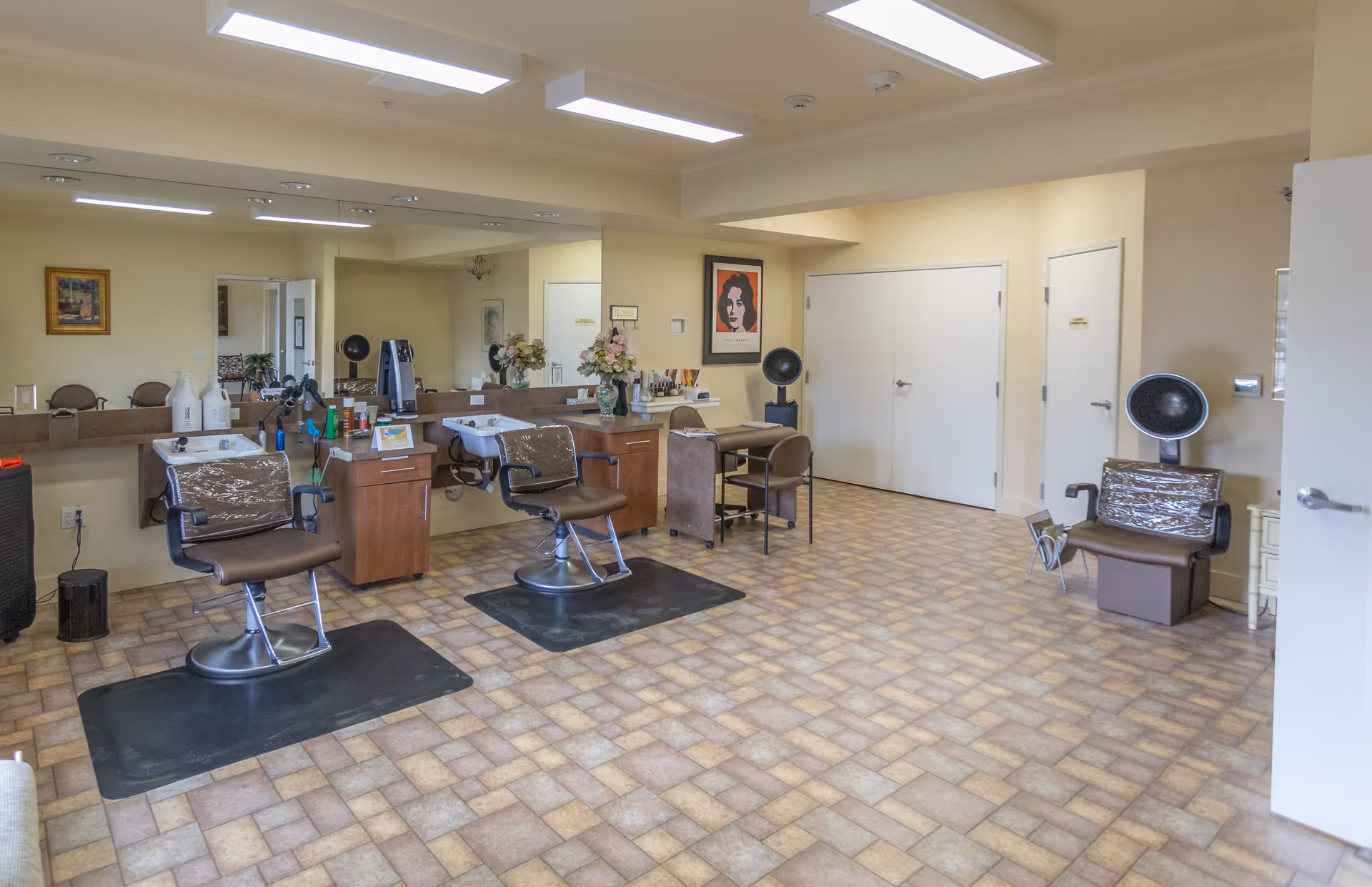 Interior view of a hair salon area in a retirement living facility with two salon chairs in front of sinks and mirrors, a hair dryer chair, a small table with chairs, and decorative flowers and artwork on the walls.