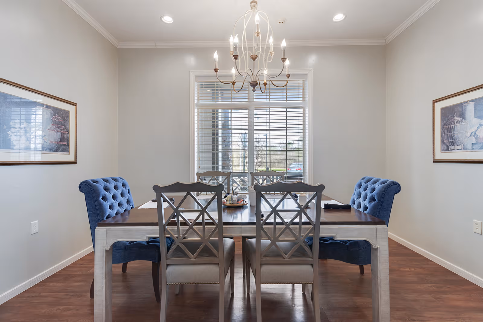 A dining room with a rectangular wooden table surrounded by six chairs, two of which are blue upholstered armchairs at the ends. The room has light gray walls, two framed artworks on either side, a large window with blinds at the far end, and a chandelier hanging above the table. The floor is wooden.