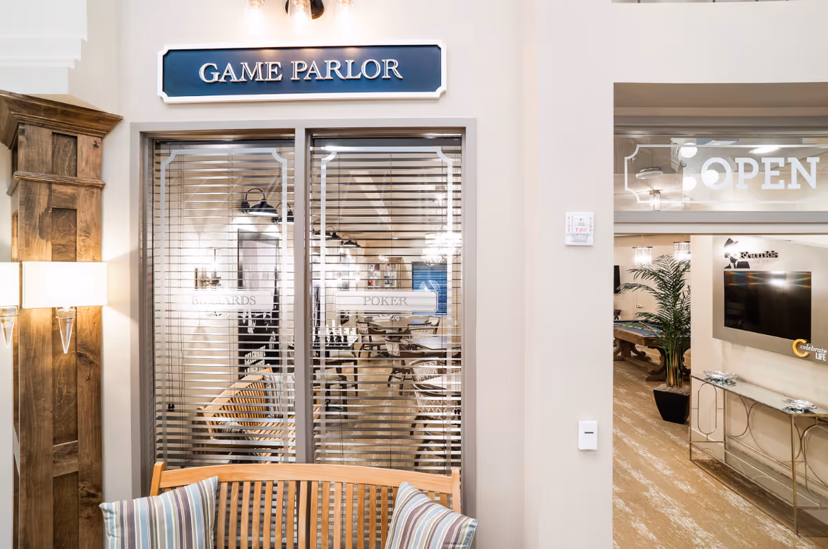 Entrance to a game parlor with glass-paneled doors and blinds, a bench with striped pillows in the foreground and a TV lounge visible to the right.
