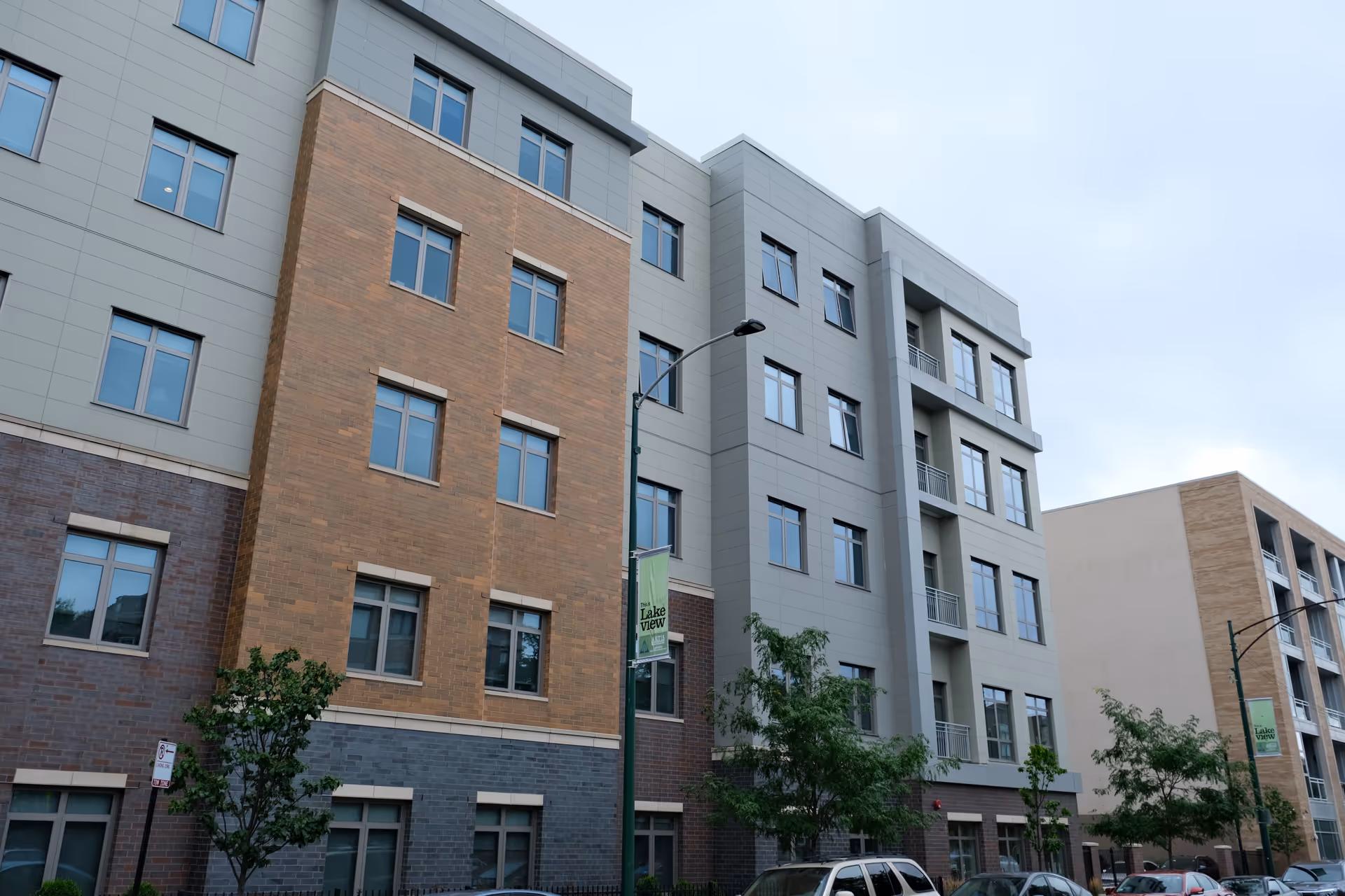 Exterior view of a modern multi-story senior living facility building with a mix of brick and panel siding, several windows, street lamps, trees, and parked cars along the street.