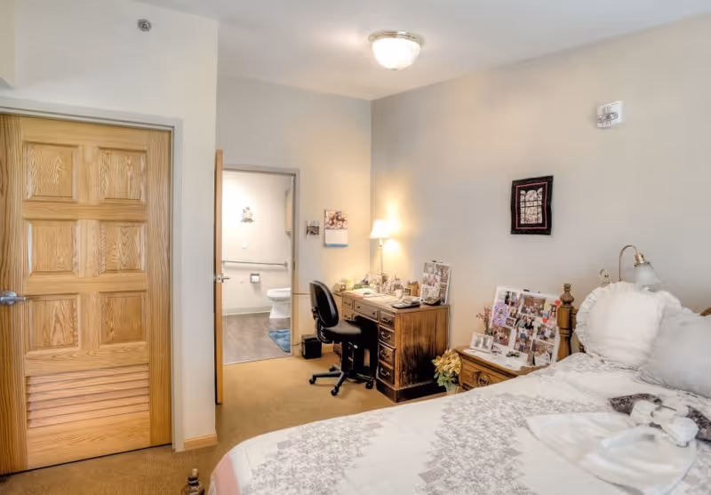 A cozy bedroom in a senior living facility featuring a bed with white and gray patterned bedding, a wooden desk with a black office chair, a small table lamp, and various personal photos and decorations. An open door reveals a bathroom with a toilet and grab bars.