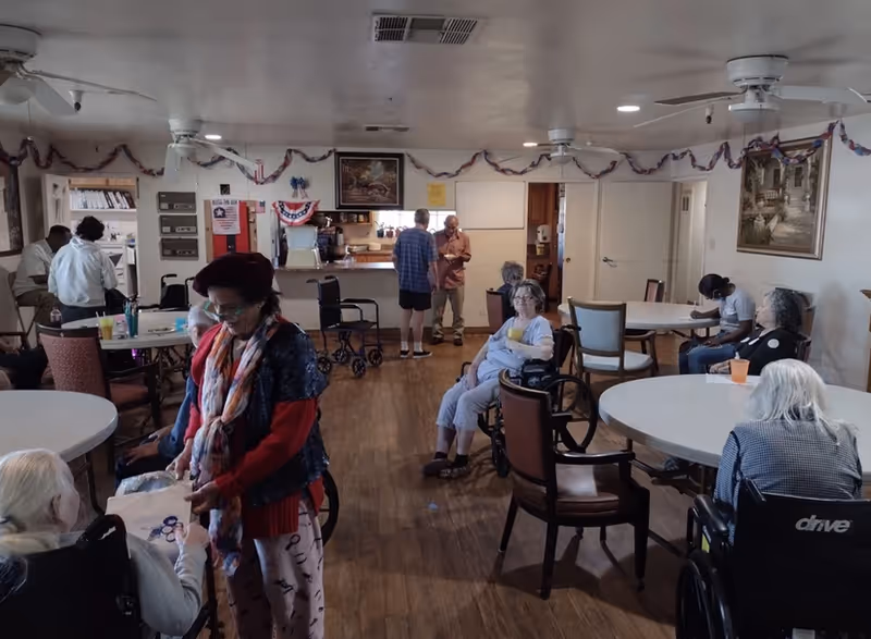 Residents sitting and socializing at tables in a communal dining/activity room of an assisted living facility.