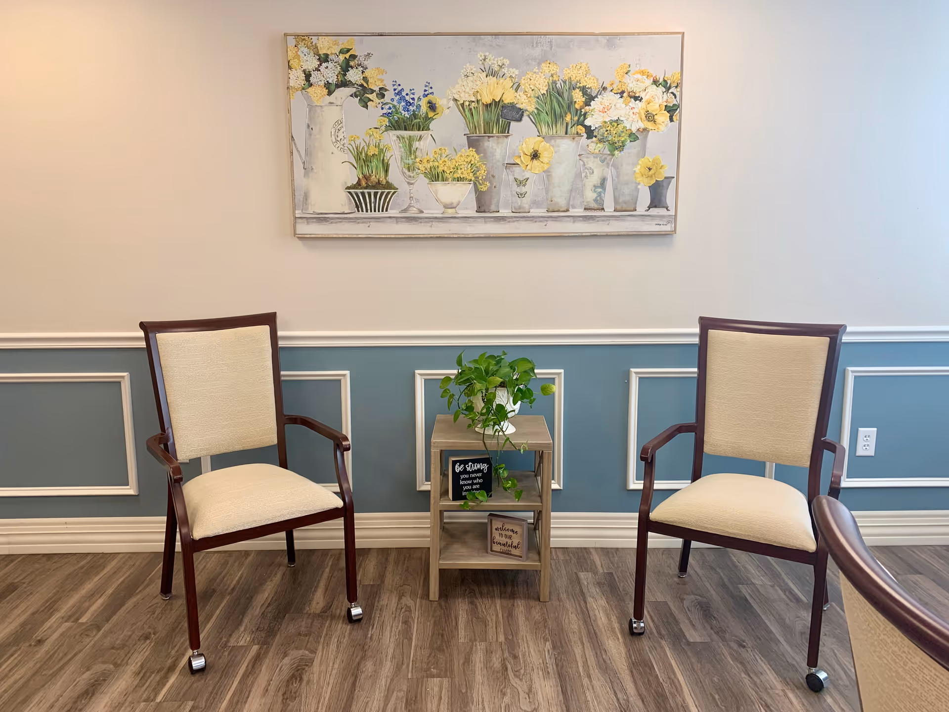Two upholstered chairs flank a small wooden side table with a potted plant beneath a floral painting on a paneled wall.