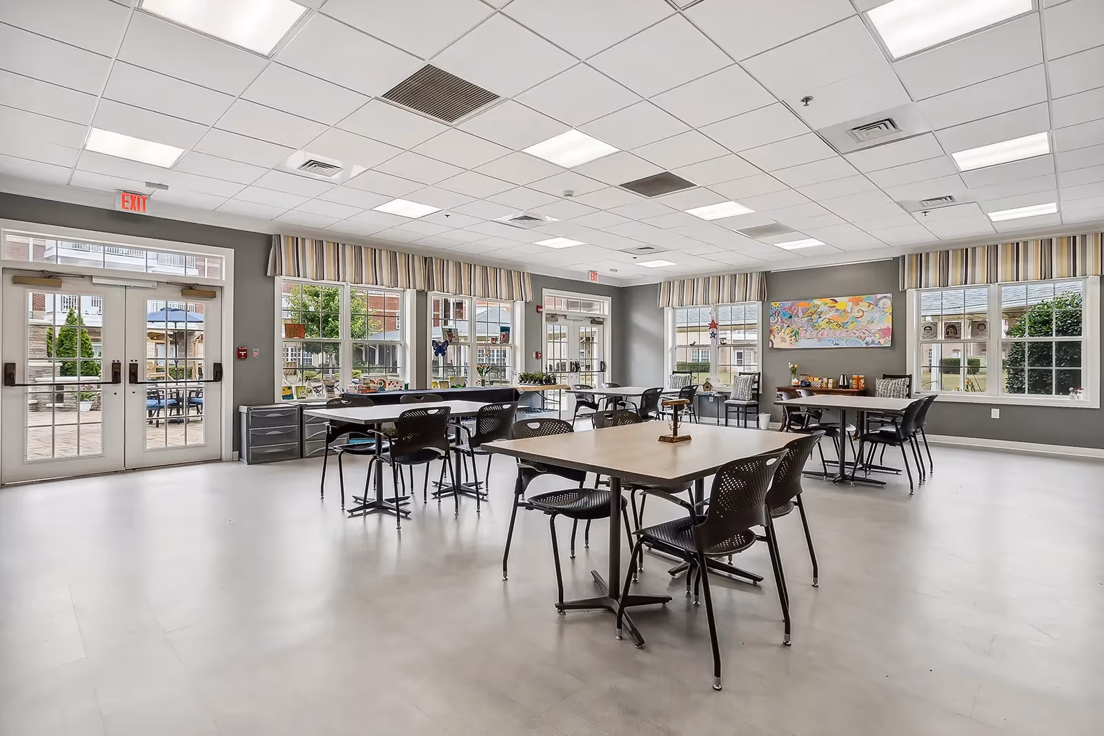 Bright multi-table dining/activity room with chairs, large windows, and double doors leading to an outdoor patio.