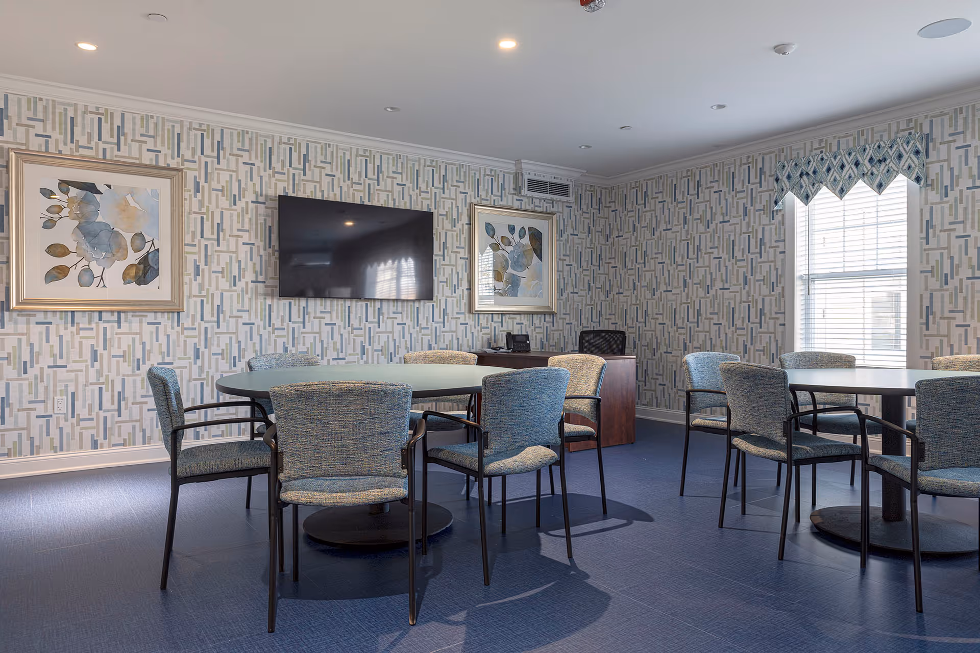 A well-lit room with two round tables surrounded by blue upholstered chairs. The walls have a geometric patterned wallpaper with two framed abstract floral paintings. A flat-screen TV is mounted on the wall between the paintings. There is a window with blinds and a valance on the right side, and a small wooden desk with a chair and a phone underneath the TV.