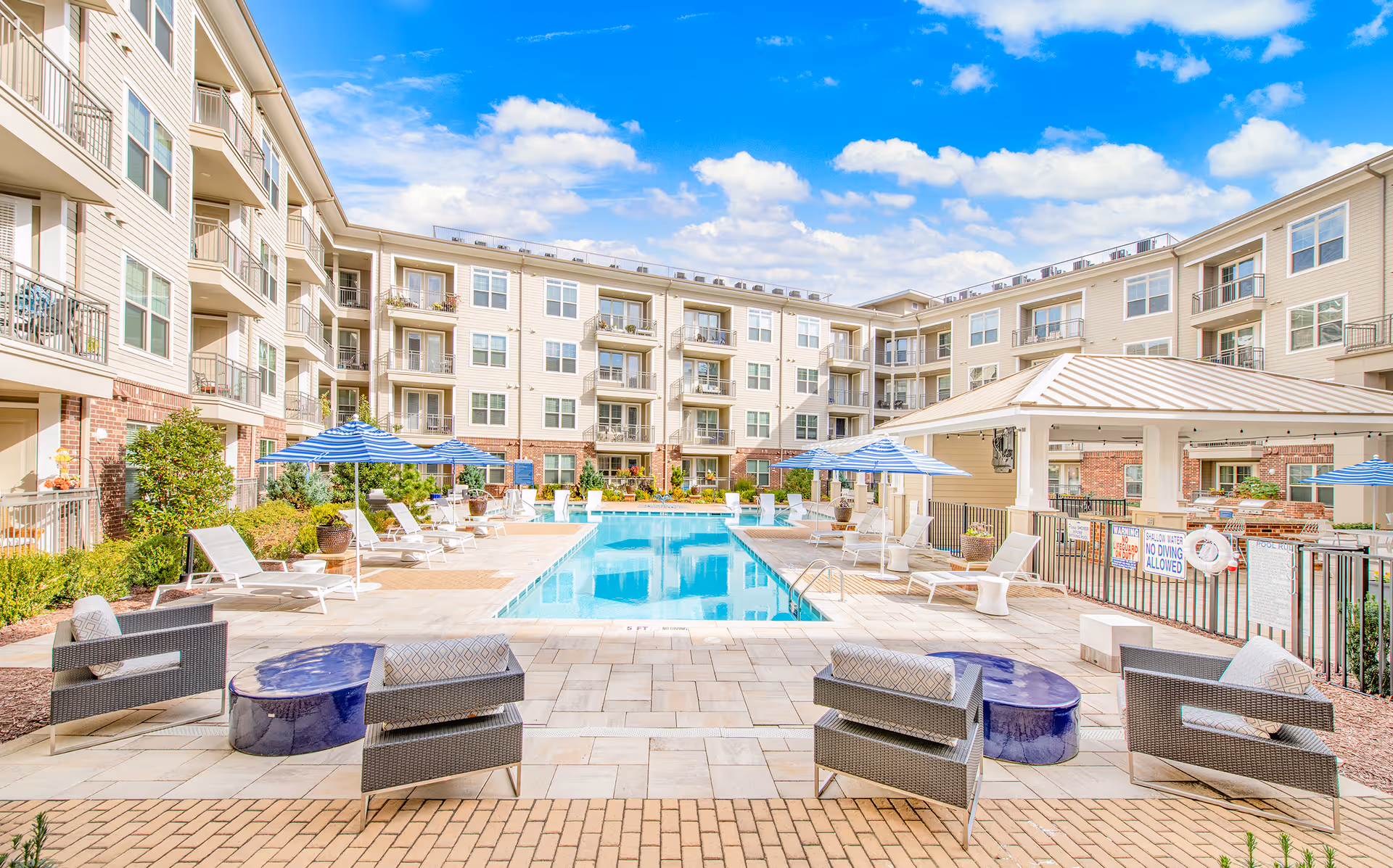 Outdoor pool area surrounded by beige apartment buildings with lounge chairs and umbrellas.
