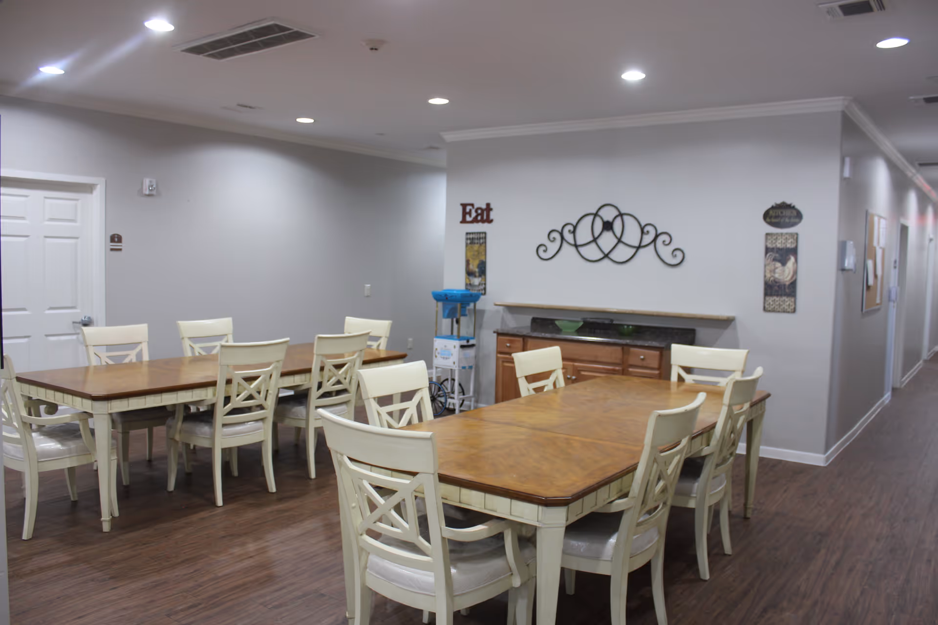 A dining area with two wooden tables and several white chairs arranged around them. The room has light gray walls, wooden flooring, and recessed ceiling lights. On the far wall, there is decorative wall art and signs, including one that says 'Eat'. A hallway extends to the right side of the image.
