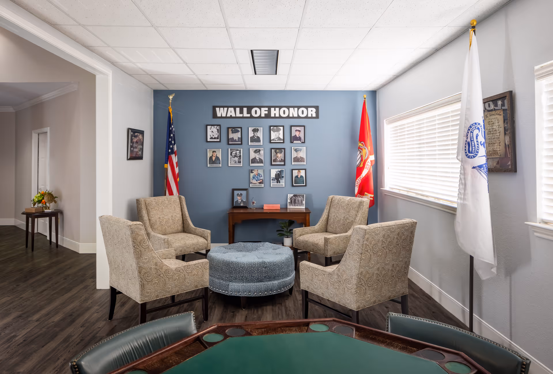 A cozy seating area with four beige patterned armchairs arranged around a round blue ottoman. Behind the chairs is a blue wall with a 'WALL OF HONOR' sign and framed photographs of military personnel. Two flags, one American and one Marine Corps, stand on either side of the wall. A window with blinds is on the right side, and a small wooden table with a plant and framed photos is beneath the wall display. The floor is dark wood, and part of a green card table is visible in the foreground.