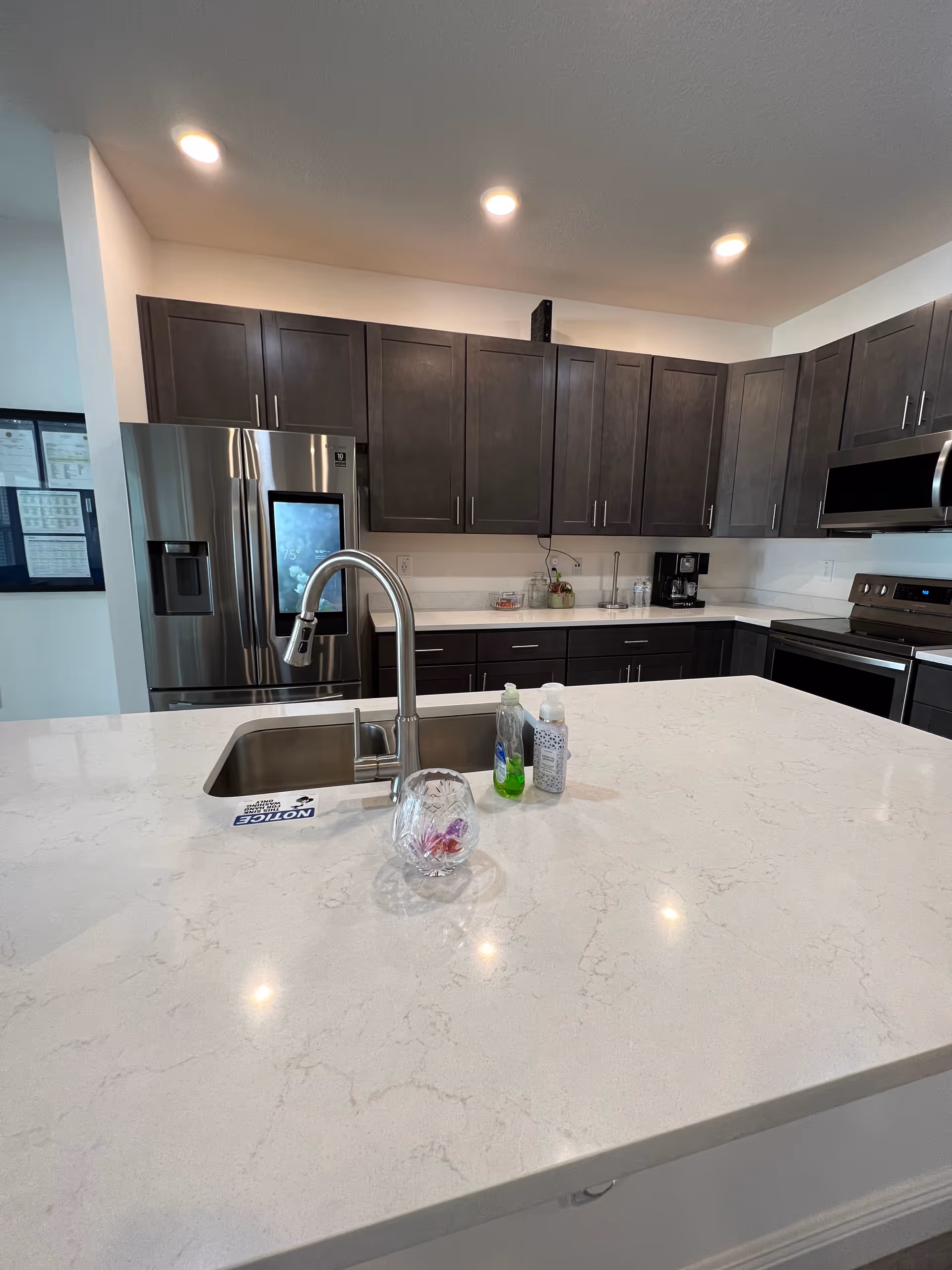 Modern kitchen with a large white island countertop, sink and faucet, stainless steel refrigerator and dark upper cabinets.