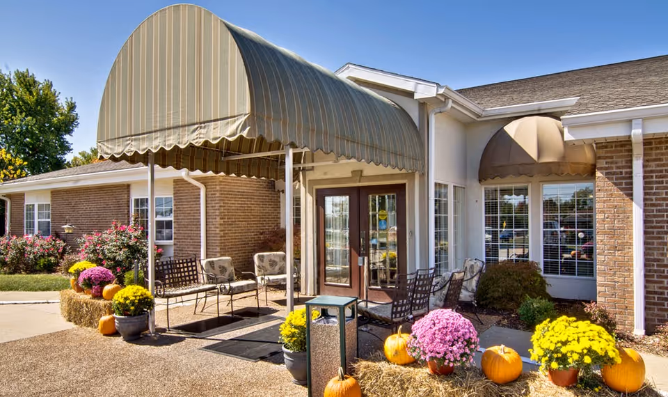 Entrance of Ashland Villa featuring a covered porch with striped awnings, outdoor seating with metal chairs and a bench, and seasonal decorations including pumpkins, hay bales, and colorful potted flowers. The building exterior is brick and light-colored siding with large windows and a glass double door.