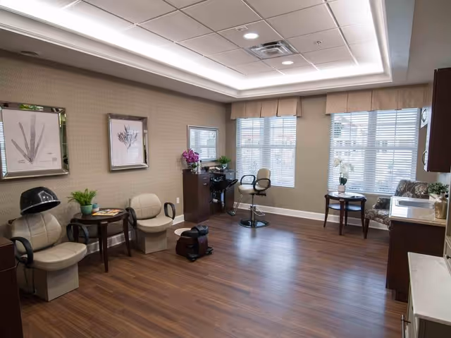 A spacious and well-lit salon room with wooden flooring and beige walls. The room contains salon chairs, a hair dryer chair, a small table with plants and magazines, a mirror, and cabinets. Large windows with blinds allow natural light to fill the space.