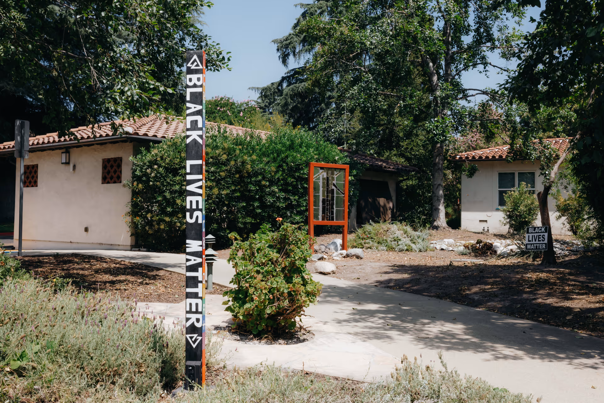 Courtyard with bungalow-style buildings, a walkway, and a vertical 'Black Lives Matter' signpost among trees and shrubs.