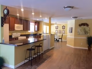 Interior view of a senior living facility showing a kitchen area with a countertop and two bar stools, a dining area with a table and chairs in the background, and a hallway with wooden flooring and wall decorations.